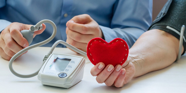 Eine Person misst den Blutdruck einer anderen, die ein rotes Stoffherz in der Hand hält.