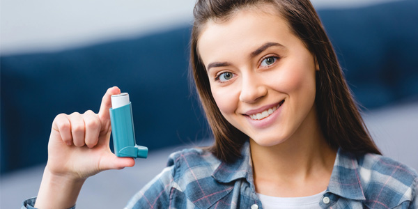Person hält einen blauen Asthmaspray-Inhalator in der rechten Hand und trägt ein kariertes Hemd.